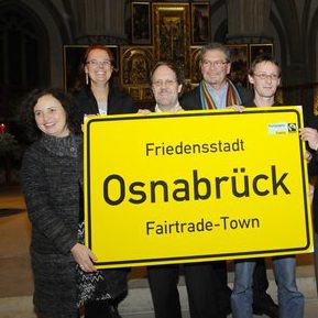 Eine Personengruppe in einer Kirchemit einem Ortseingangsschild in Originalgröße in den Händen mit der Aufschrift "Friedensstadt Osnabrück. Fairtrade-Town"