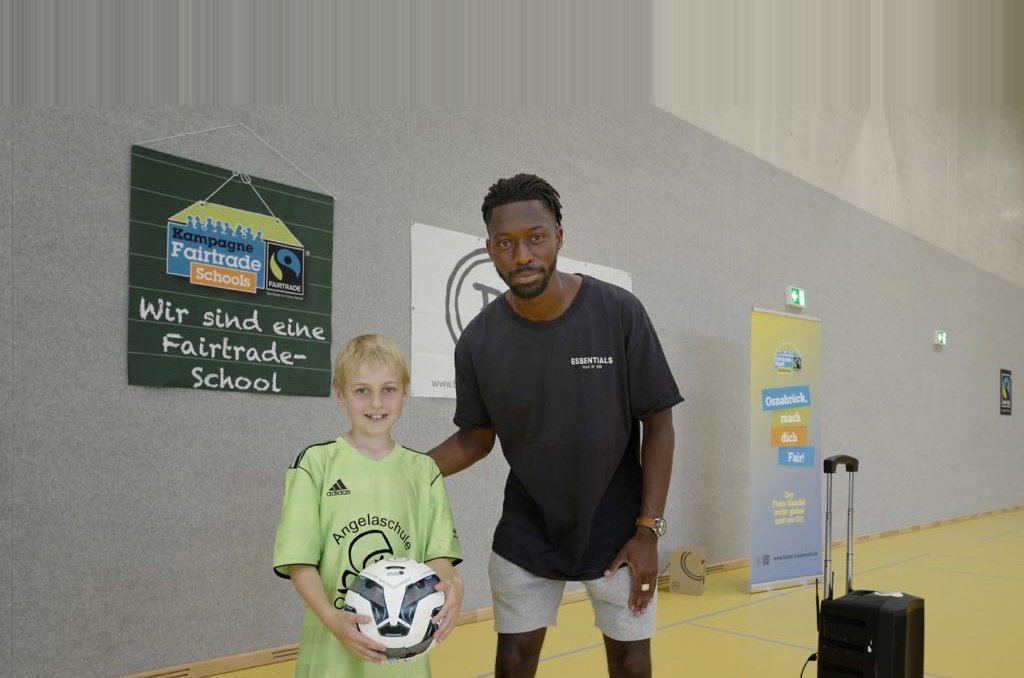 Ein Junge mit einem grünen Sportshirt und einem Fußball neben einem dunkelhäutigen Erwachsenen in Sportkleidung
