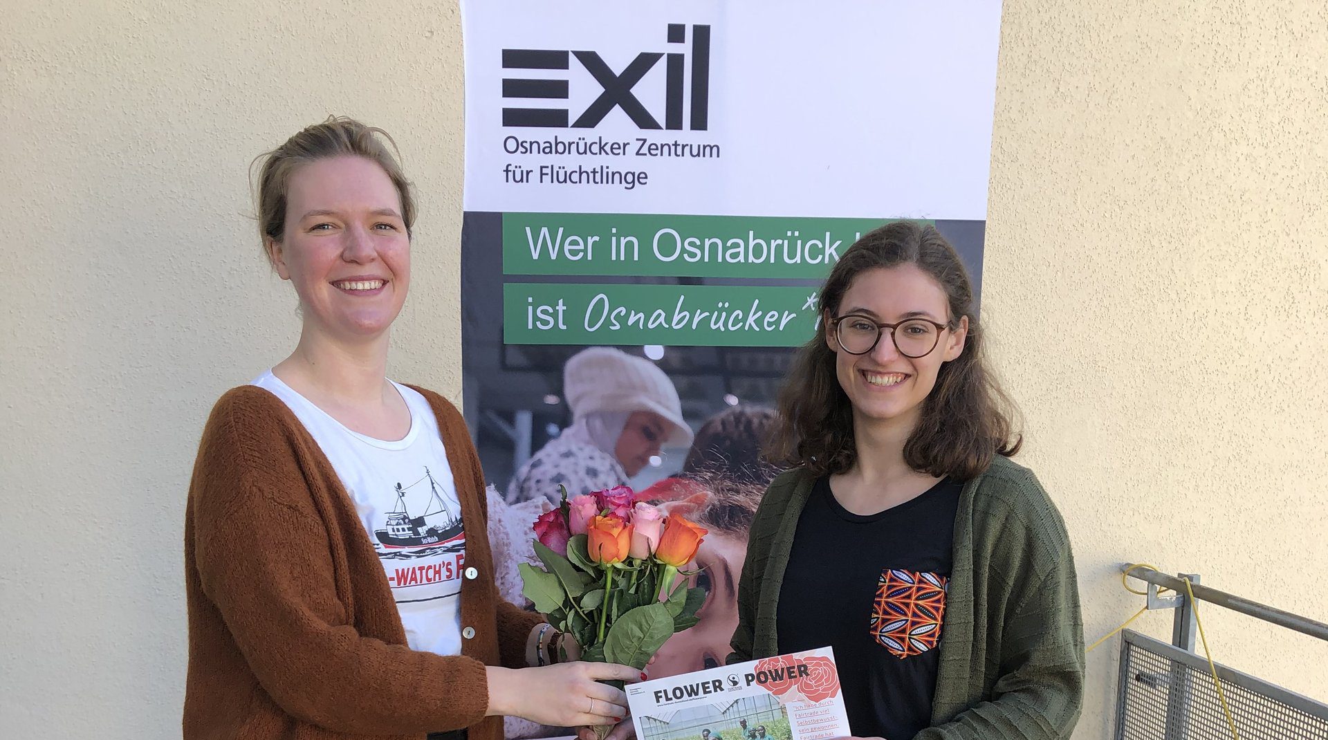 Eine Frau gratuliert einer anderen Frau mit Blumen, im Hintergrund ein Roll-Up mit der Aufschrift "Exil"