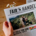 Einer Person wird über die Schulter geschaut, während sie eine Zeitung mit dem Titel "Fair Handeln" ließt. Das Titelbild zeigt eine Frau mit Hut, die ein beladenes Maultier in einer Berglandschaft führt.